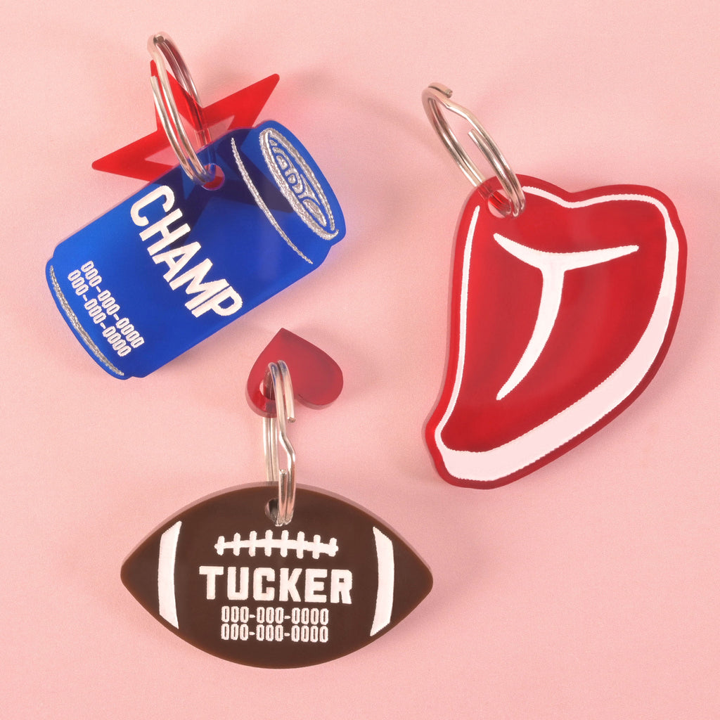 A collection of three personalized pet tags. A blue soda can-shaped tag says "CHAMP" and has a red star charm. A brown football-shaped tag says "TUCKER" and has a small red heart charm. A red steak-shaped tag is also shown. All tags have placeholder phone numbers and are attached to a silver ring for a collar. The items are displayed on a solid pink background.