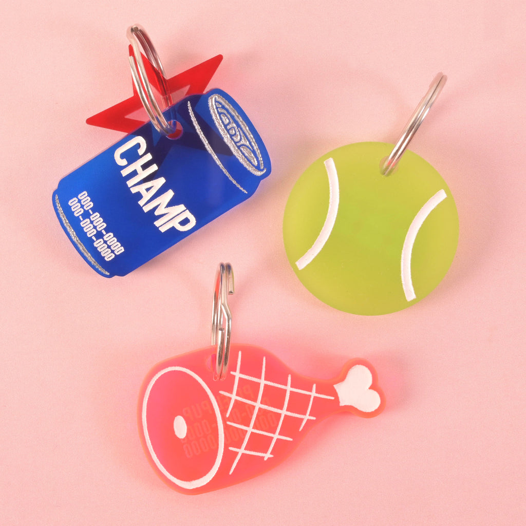 A collection of three personalized pet tags. A blue soda can-shaped tag says "CHAMP" and has a red star charm. A light green tennis ball-shaped tag is also shown. A pink ham-shaped tag is also visible. All tags have placeholder phone numbers and are attached to a silver ring for a collar. The items are displayed on a solid pink background.
