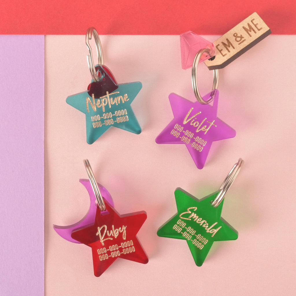 A collection of four personalized star and moon-shaped pet tags in various colors on a background of pink, red, and purple. The tags include a translucent teal star for "Neptune," a frosted purple star for "Violet," a pink-red star for "Ruby," and a frosted green star for "Emerald." Each tag has a placeholder phone number and is attached to a silver ring for a collar. A small wooden "EM & ME" tag and a translucent pink gemstone charm are also visible.