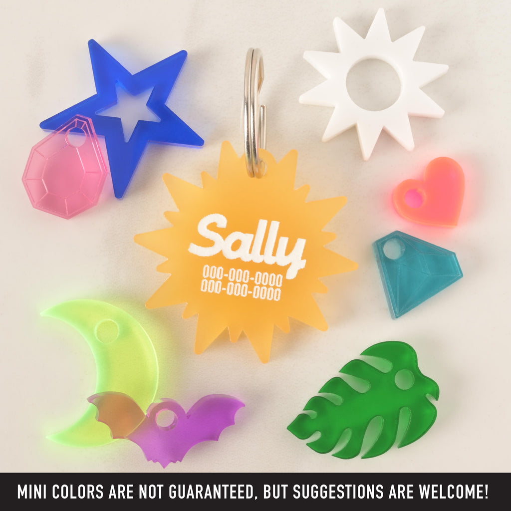 A collection of pet tag and charms scattered on a white surface. The central piece is a translucent yellow sunburst-shaped tag with the name "Sally" and a placeholder phone number. Surrounding it are various charms, including a blue star with a pink gemstone, a neon yellow moon with a purple bat, a green monstera leaf, a white sun, a neon pink heart, and a blue diamond. The image also contains the text "MINI COLORS ARE NOT GUARANTEED, BUT SUGGESTIONS ARE WELCOME!"