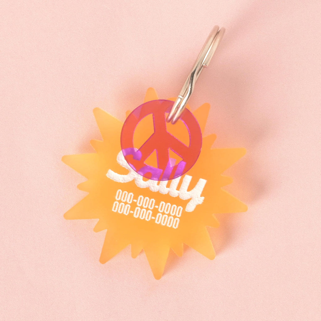 A custom, orange, sun-shaped pet ID tag. The tag is engraved with the name "Sally" and a phone number in white font. A translucent purple peace sign charm is attached, sitting in front of the main tag. Both are on a shiny silver keyring and are displayed on a solid pink background.