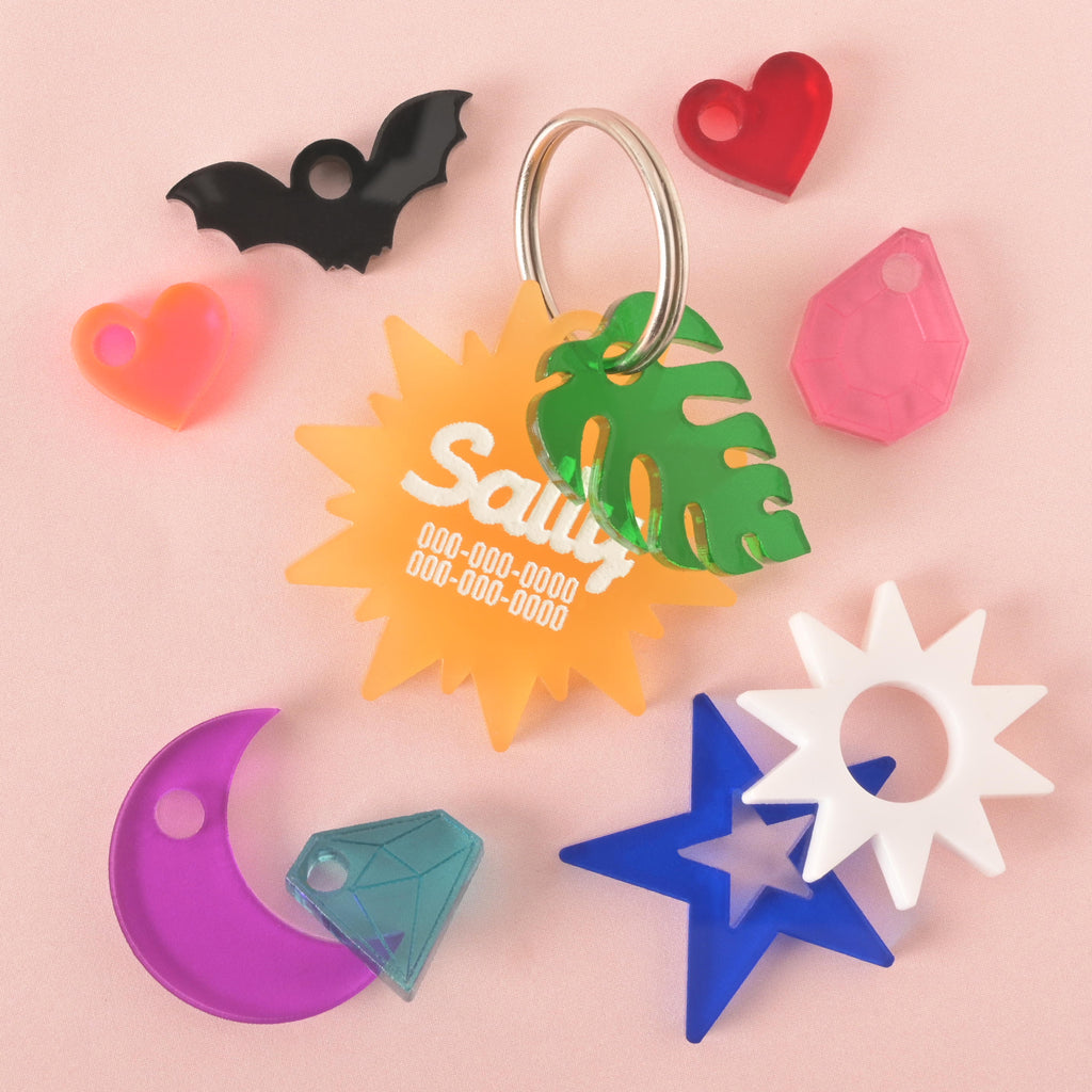 A collection of pet tags and charms scattered on a light pink background. The central piece is a translucent orange sunburst-shaped tag with the name "Sally" and a placeholder phone number. Surrounding it are various charms, including a black bat, a green monstera leaf, a white sun, a blue star with a blue diamond, a neon pink heart, a pink gemstone, and a purple moon.