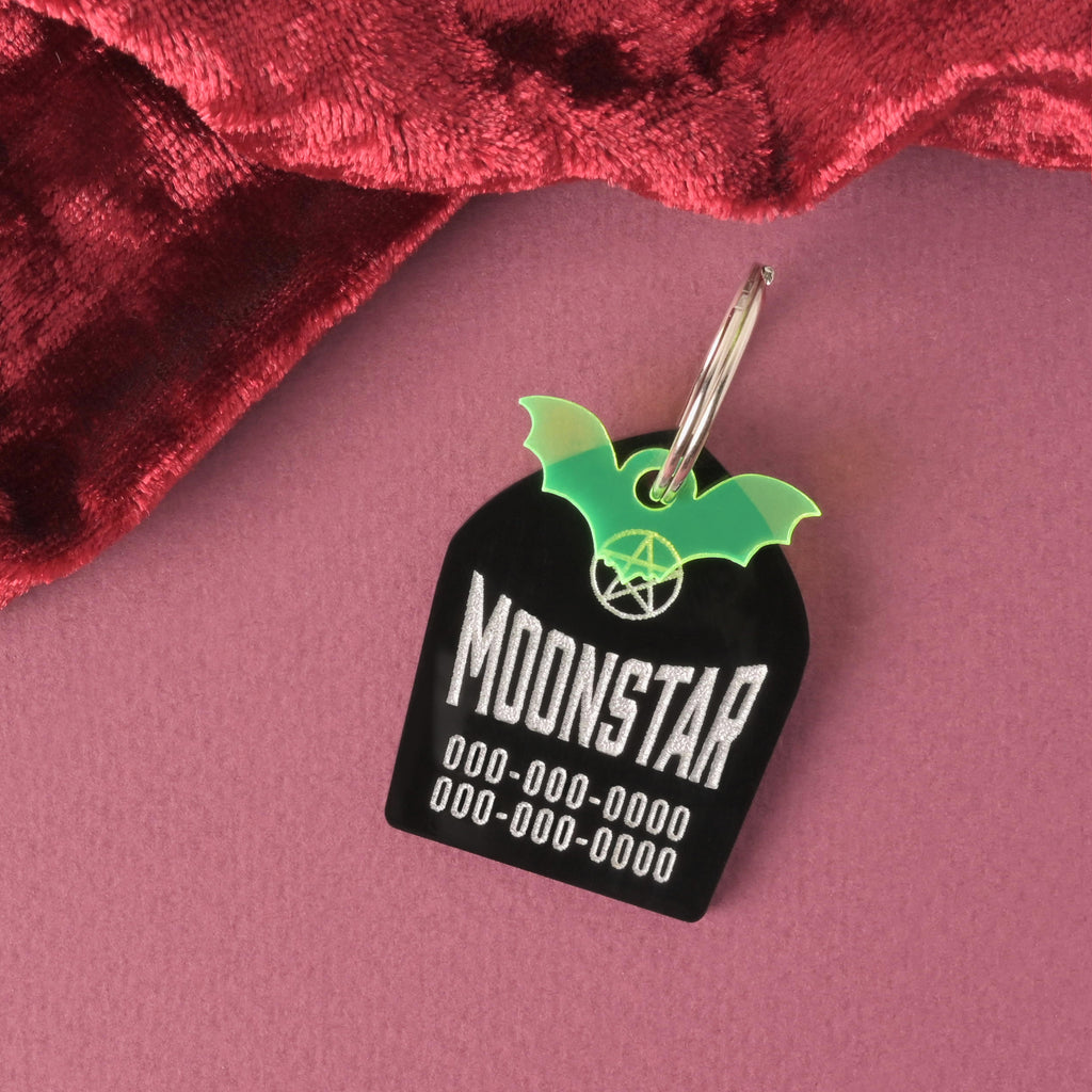 A personalized black tombstone-shaped pet ID tag for a dog or cat. The tag is engraved with the name 'MOONSTAR' and three lines of phone numbers, featuring a white pentagram graphic and a neon green glow-in-the-dark bat charm attached to the ring.