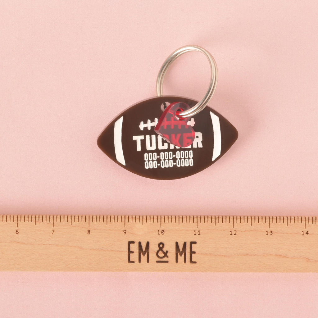 A personalized brown football-shaped pet tag with the name "TUCKER" and a placeholder phone number on it. A small, transparent red heart charm is attached behind the tag. A wooden ruler with the "EM & ME" logo is placed below the tag to show its size. The items are displayed on a solid pink background.