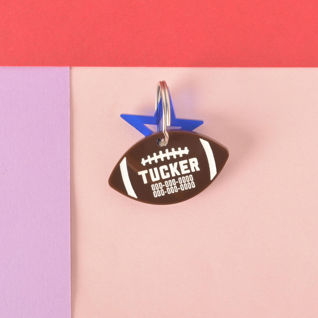 A personalized brown football-shaped pet tag with the name "TUCKER" and a placeholder phone number. The tag has a small, translucent blue star charm attached. Both are connected to a silver ring, perfect for a collar, and are displayed on a background of solid pink, purple, and red.