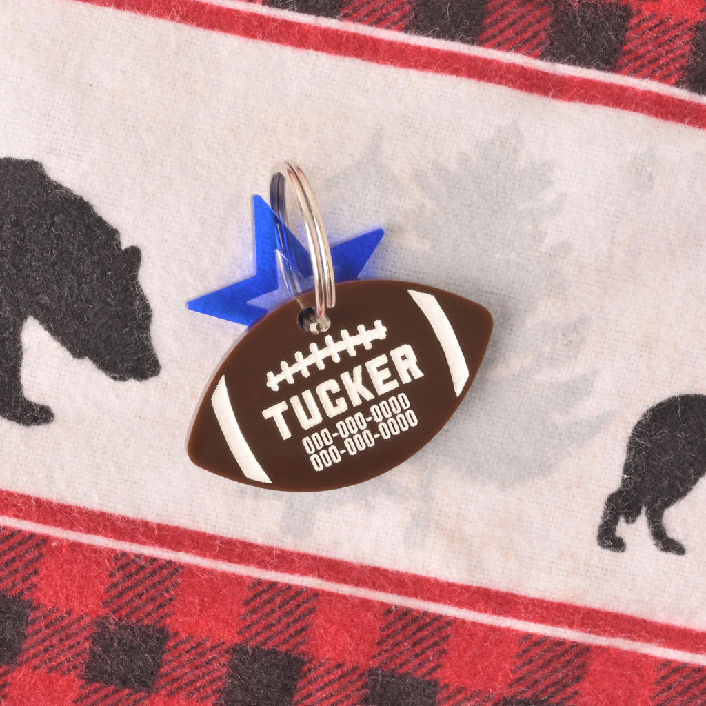 A personalized brown football-shaped pet tag with the name "TUCKER" and a placeholder phone number. The tag has a small, translucent blue star charm attached. Both are connected to a silver ring, perfect for a collar, and are displayed on a red and black flannel blanket with a white and red border and a black bear print.