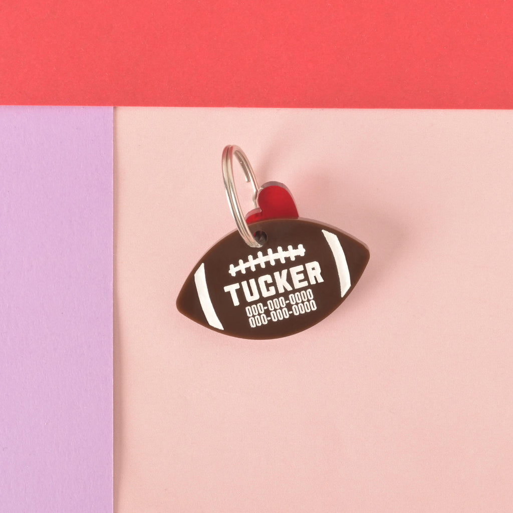 A personalized brown football-shaped pet tag with the name "TUCKER" and a placeholder phone number on it. A small red heart charm is attached to the tag. Both are connected to a silver ring for a collar and are displayed on a light pink, purple, and red background.