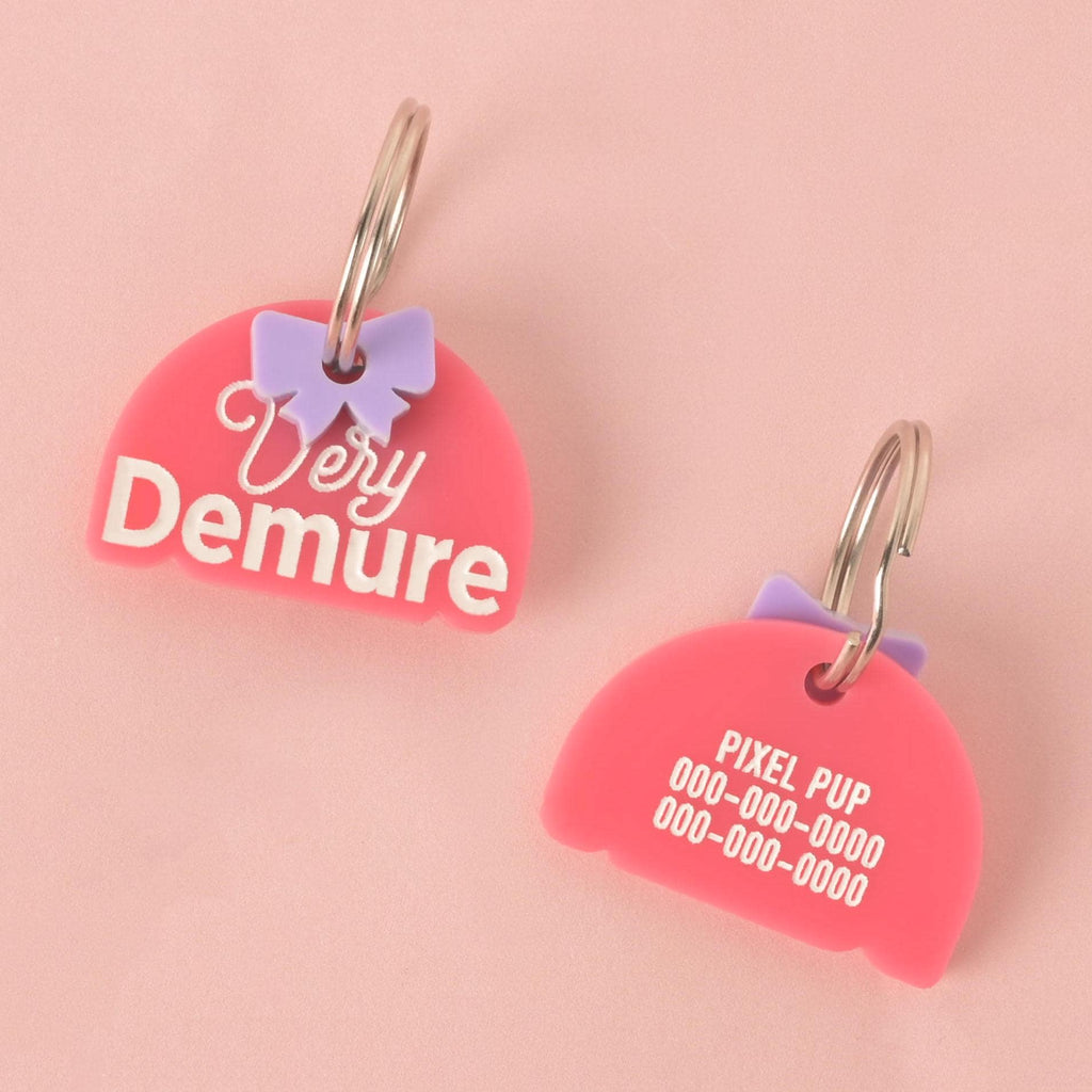 A pair of custom, half-moon-shaped pet ID tags in bright pink. Both tags have a small, light purple bow charm attached to a silver keyring. The tag on the left is engraved with "Very Demure." The tag on the right is engraved with "PIXEL PUP" and a phone number. They are displayed on a solid pink background.