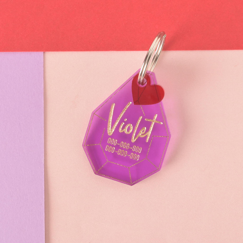 A personalized purple gemstone-shaped pet tag with the name "Violet" written in a gold-colored script, along with a placeholder phone number. A small red heart charm is attached to the tag. Both are connected to a silver ring for a collar and are displayed on a light pink, purple, and red background.