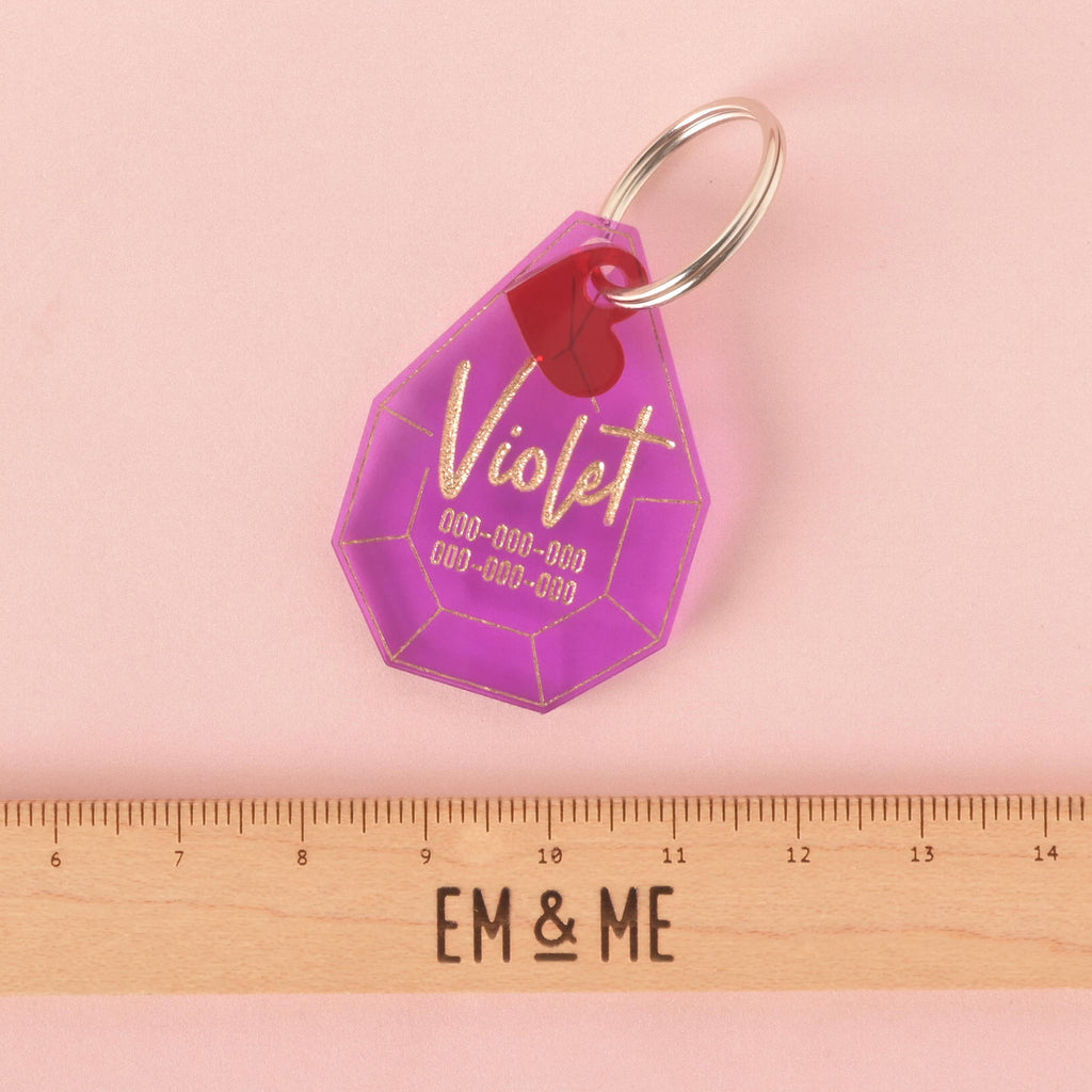 A personalized purple gemstone-shaped pet tag with the name "Violet" written in a gold-colored script. A small red heart charm is attached. A wooden ruler with the "EM & ME" logo is placed below the tag to show its size. The tag is attached to a silver ring for a collar and is displayed on a solid pink background.