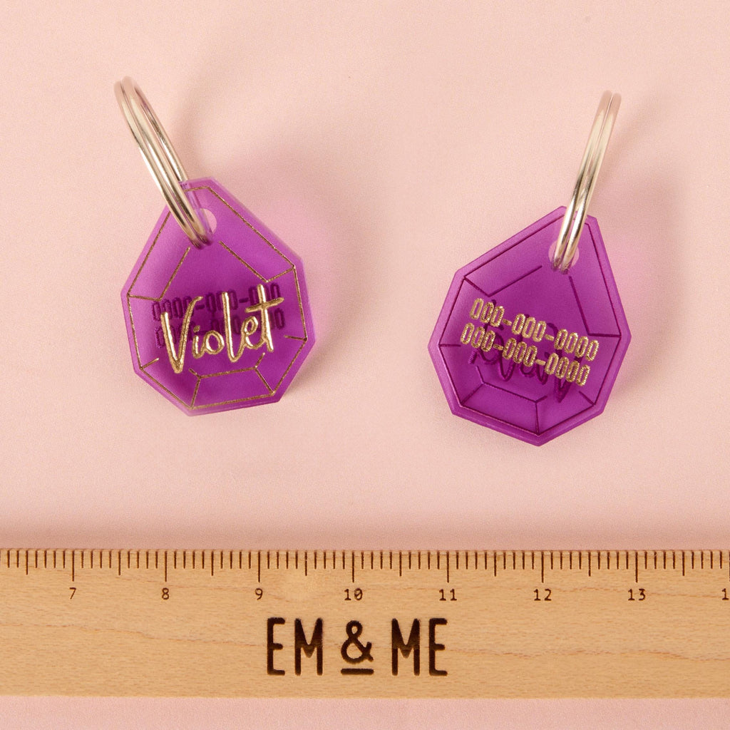 A pair of personalized purple gemstone-shaped pet tags with a ruler for scale. The tag on the left has the name "Violet" written in a gold-colored script, while the back of the tag on the right has a placeholder phone number. The tags are connected to a silver ring and are displayed on a solid pink background.