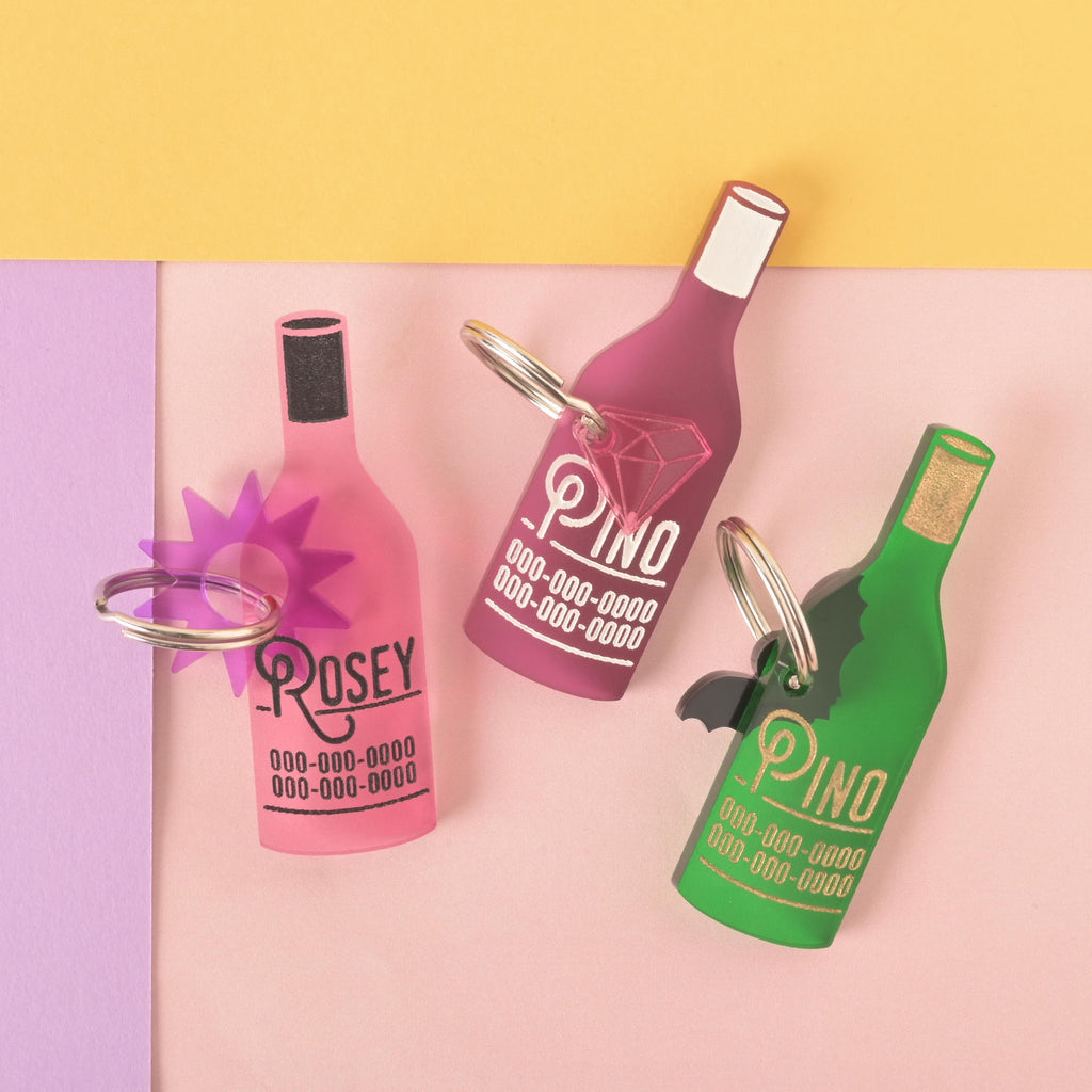 A collection of three personalized wine bottle-shaped pet tags in different colors on a background of solid pink, purple, and yellow. The tags include a pink one for "ROSEY," a dark pink one for "PINO," and a green one for "PINO." Each tag has a placeholder phone number and is attached to a silver ring for a collar. The items are also shown with small charms like a purple starburst, a pink diamond, and a black bat.