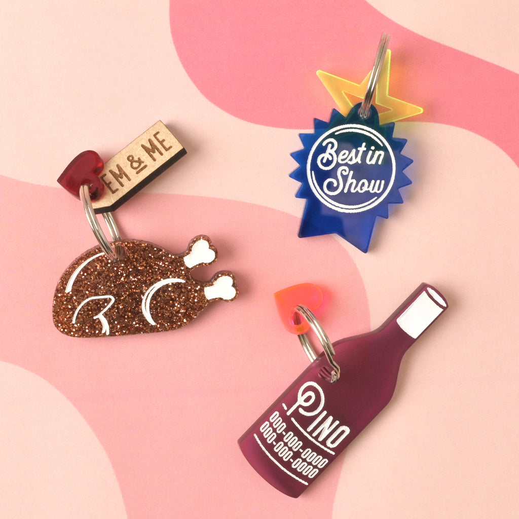 A collection of three personalized pet tags and a charm. The tags include a glittery copper turkey leg, a purple wine bottle that says "Pino," and a blue ribbon that says "Best in Show." The tags are shown with small charms like a red heart and a yellow starburst, and a small wooden "EM & ME" tag is attached to the turkey leg. The items are displayed on a light pink and solid pink background with a wavy design.