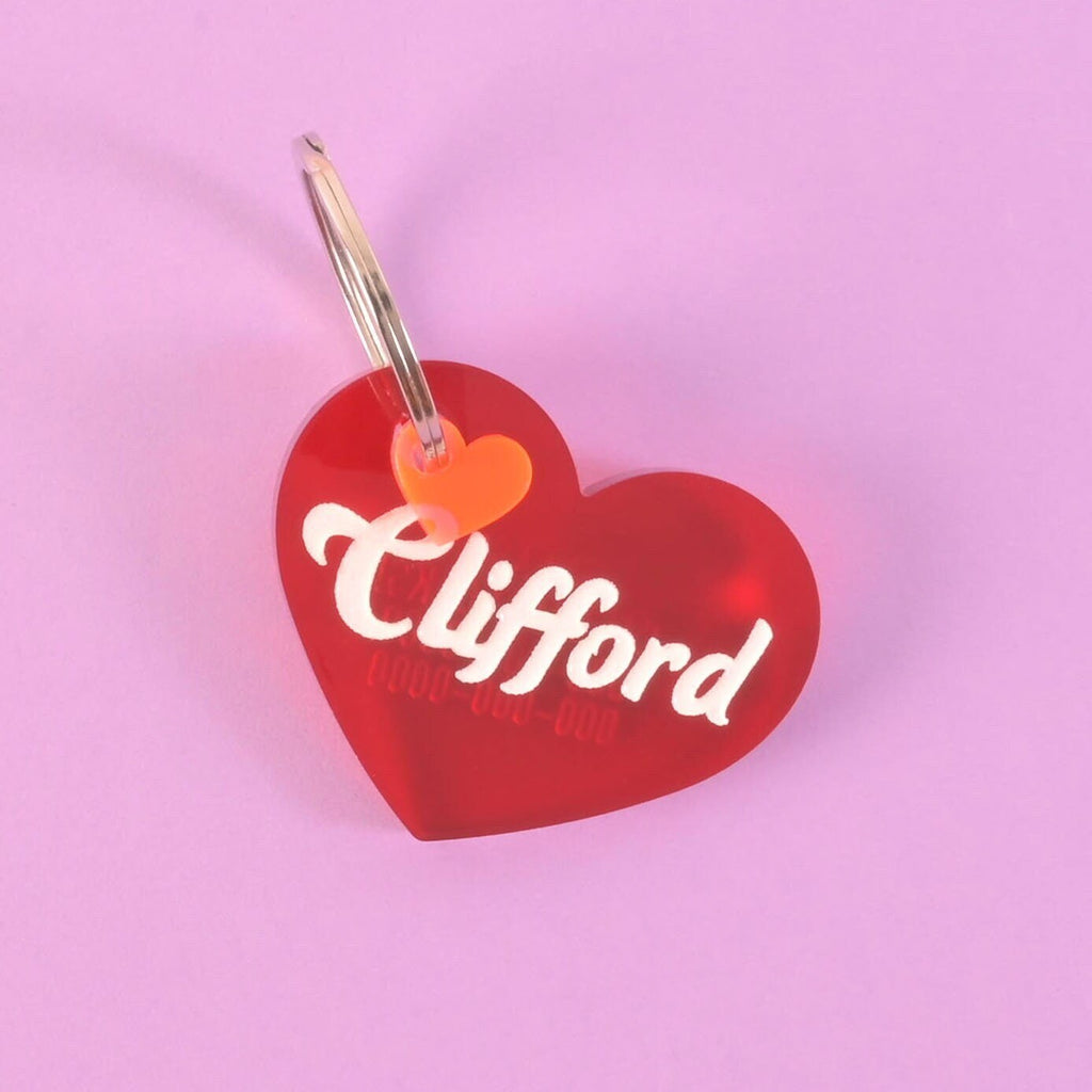 A personalized red heart-shaped pet tag with the name "Clifford" and a placeholder phone number on it. A small fluorescent pink heart charm is attached. Both are connected to a silver ring, perfect for a collar, and are displayed on a solid purple background.