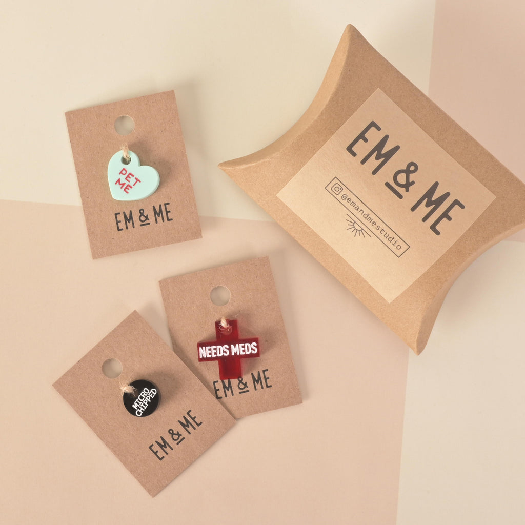 A collection of pet-themed charms and gifts on a solid beige background. The items include a small heart-shaped charm that says "PET ME," a red cross that says "NEEDS MEDS," and a black button that says "MICROCHIPPED." The charms are displayed on brown paper cards that read "EM & ME." A brown box with a black "EM & ME" logo is also shown.