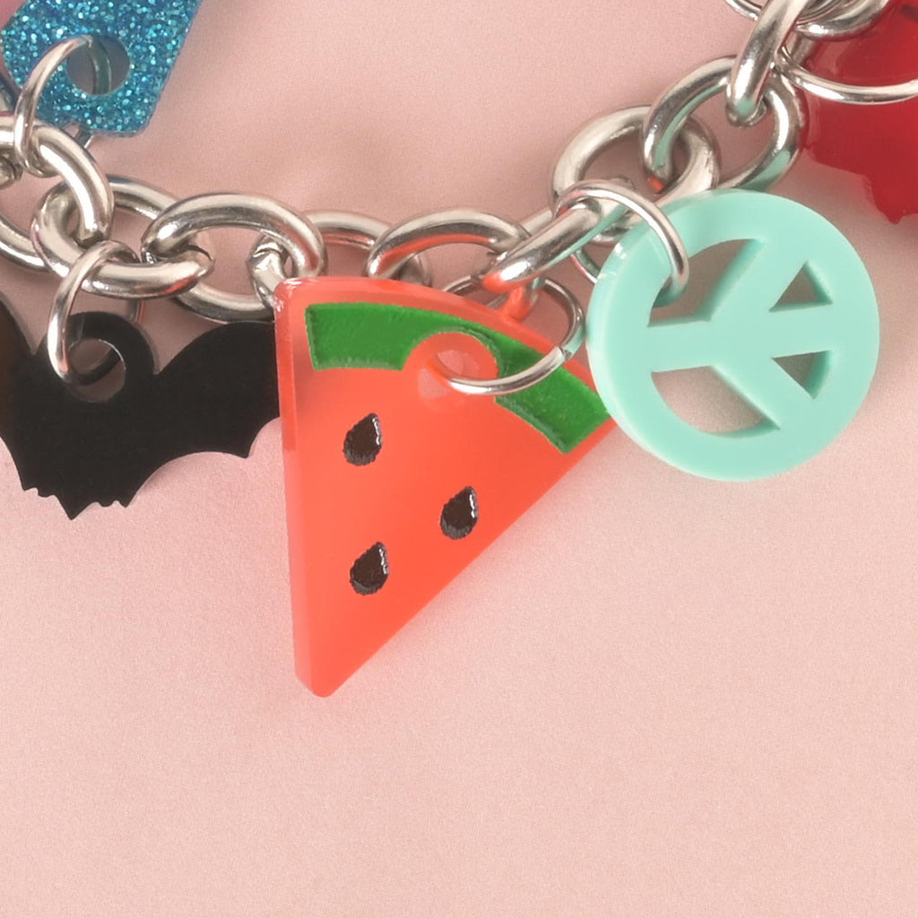 A close-up shot of a thick silver chain with several charms attached. The charms include a black bat, a pink and green watermelon slice with black seeds, and a mint green peace sign. The charms and chain are displayed on a light pink background.