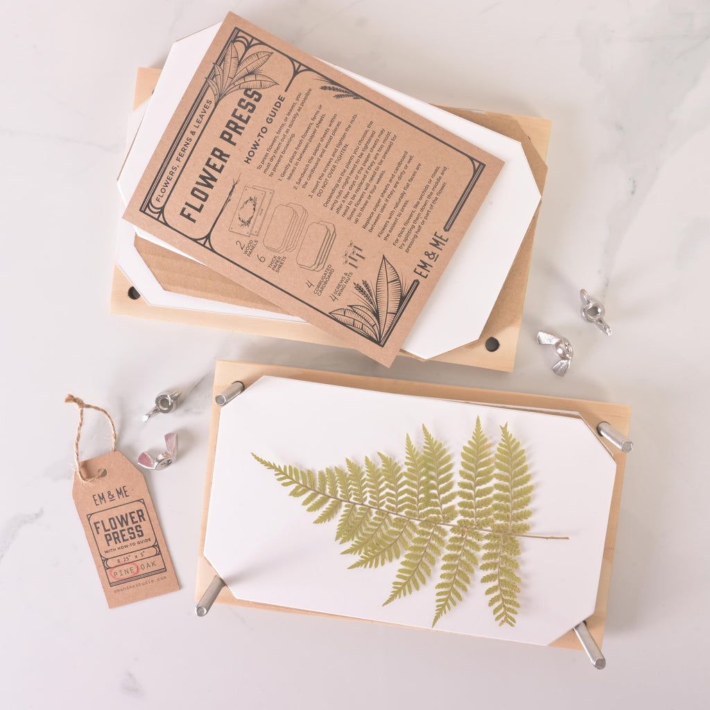 Open wooden flower press showing a pressed green fern leaf with paper sheets and instruction guide, surrounded by wing nuts and a kraft tag labeled “Flower Press.”