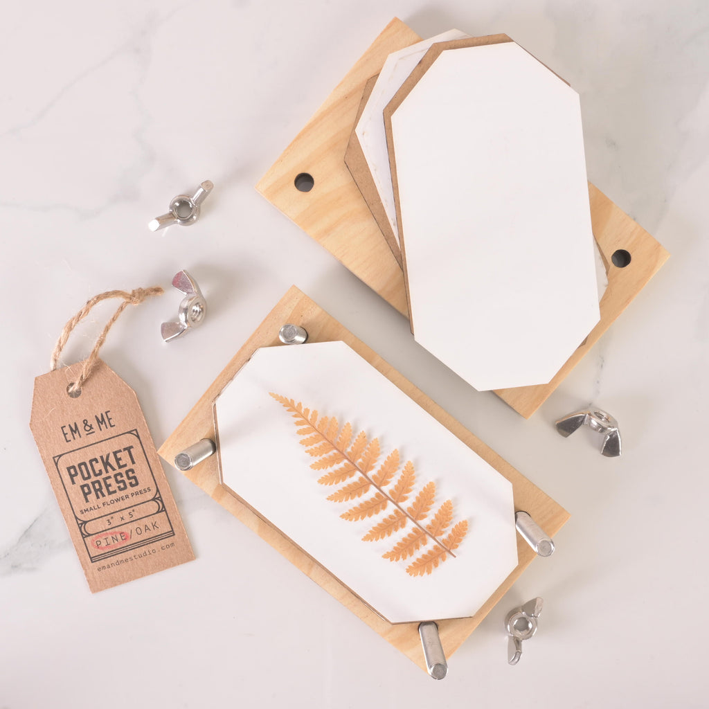 Small wooden pocket flower press with layered blotting sheets, metal wing nuts, and kraft tag, shown pressing a dried fern leaf for botanical crafts.