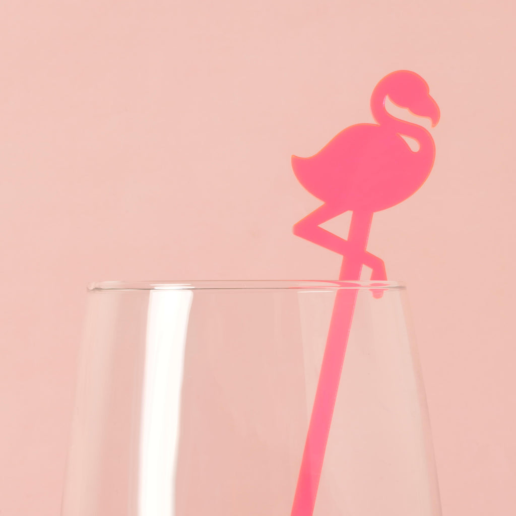 A single pink flamingo-shaped cocktail stirrer is placed inside a clear glass. The background is a solid pink color.