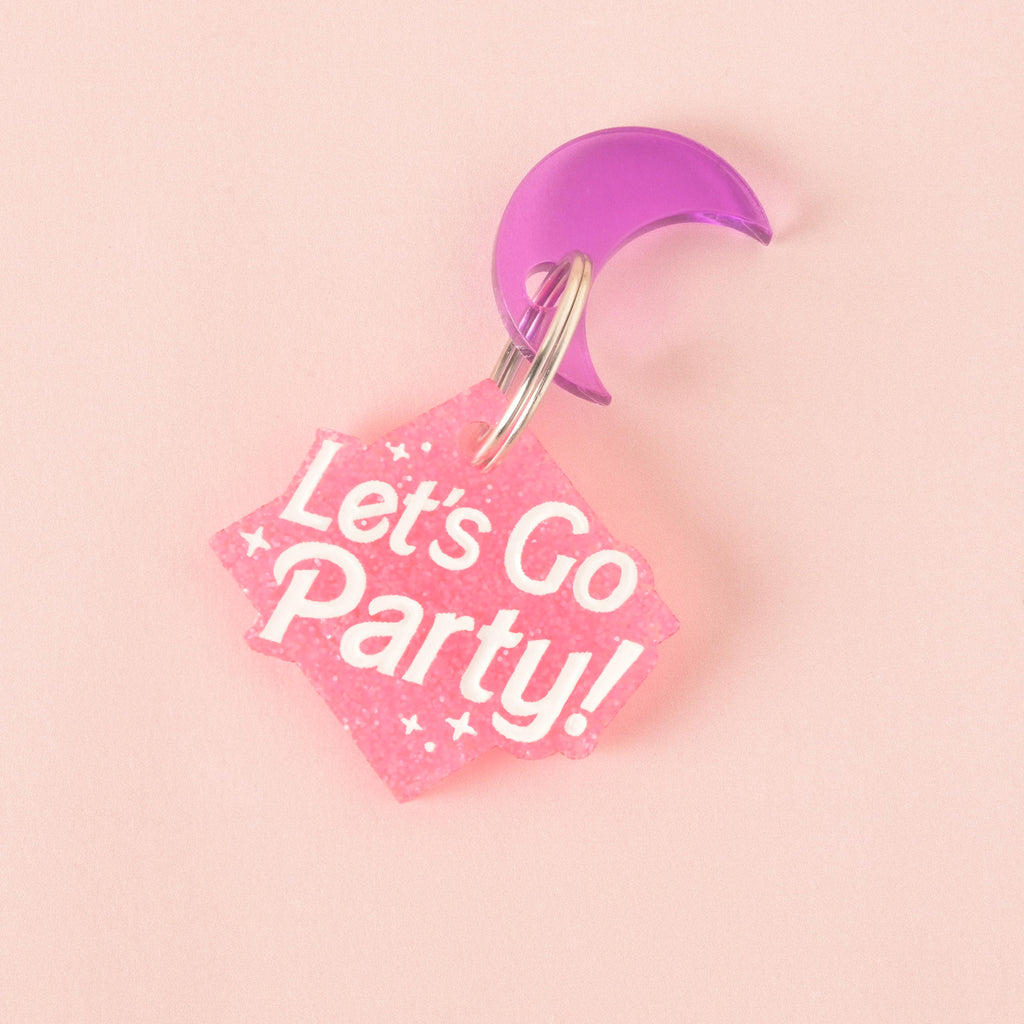 A close-up product photo of a pink glittery, rhombus-shaped pet tag with the words "Let's Go Party!" engraved on it. A purple crescent moon charm is attached to the top of the tag, which is shown with a silver split key ring.