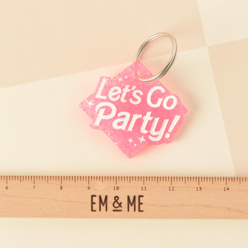 A close-up product photo of a pink glittery, rhombus-shaped pet tag with the words "Let's Go Party!" engraved on it. The tag is shown with a silver split key ring, and a wooden ruler with the words "EM & ME" is positioned below to show its size.