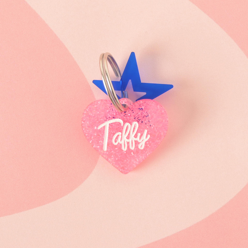 A close-up product photo of a pink glittery, heart-shaped pet tag with the name "Taffy" engraved on it. A blue star charm is attached to the top of the tag, which is shown with a silver split key ring.