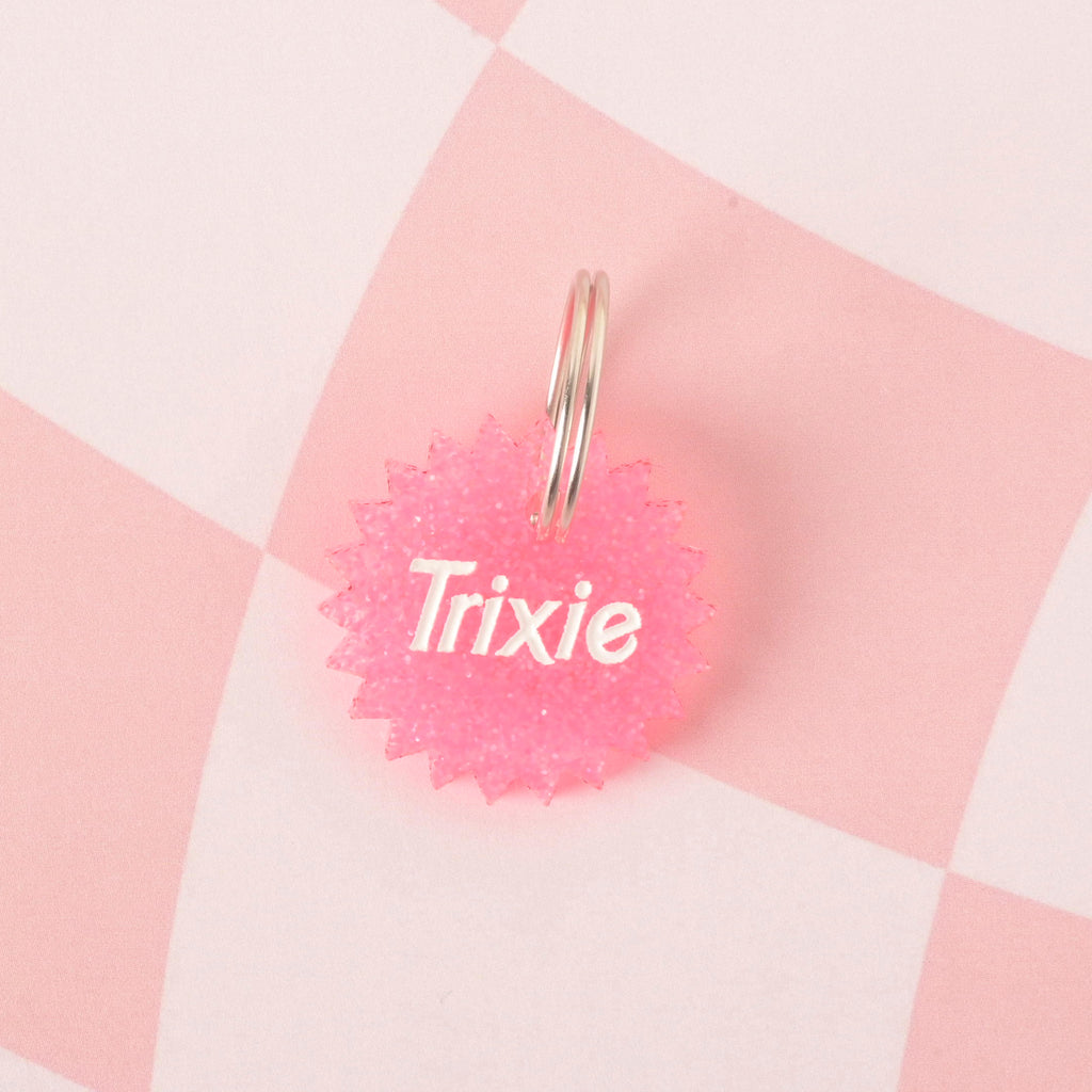A close-up product photo of a round, pink glittery pet ID tag with a unique starburst or spiked edge. The tag is engraved with the name "Trixie" and is shown with a silver split key ring on a checkered pink and white background.