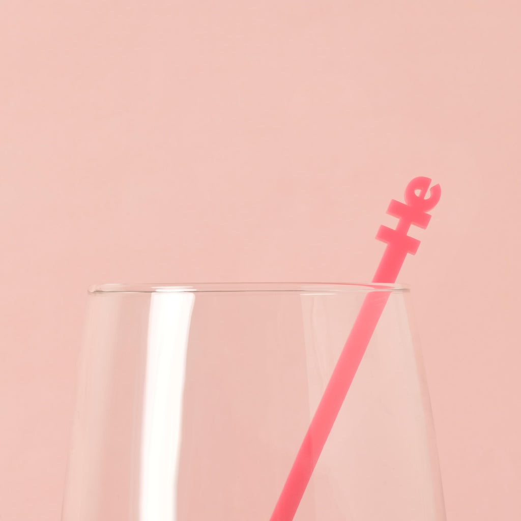 A single pink cocktail stirrer with the word "he" is placed inside a clear glass. The background is a solid pink color.