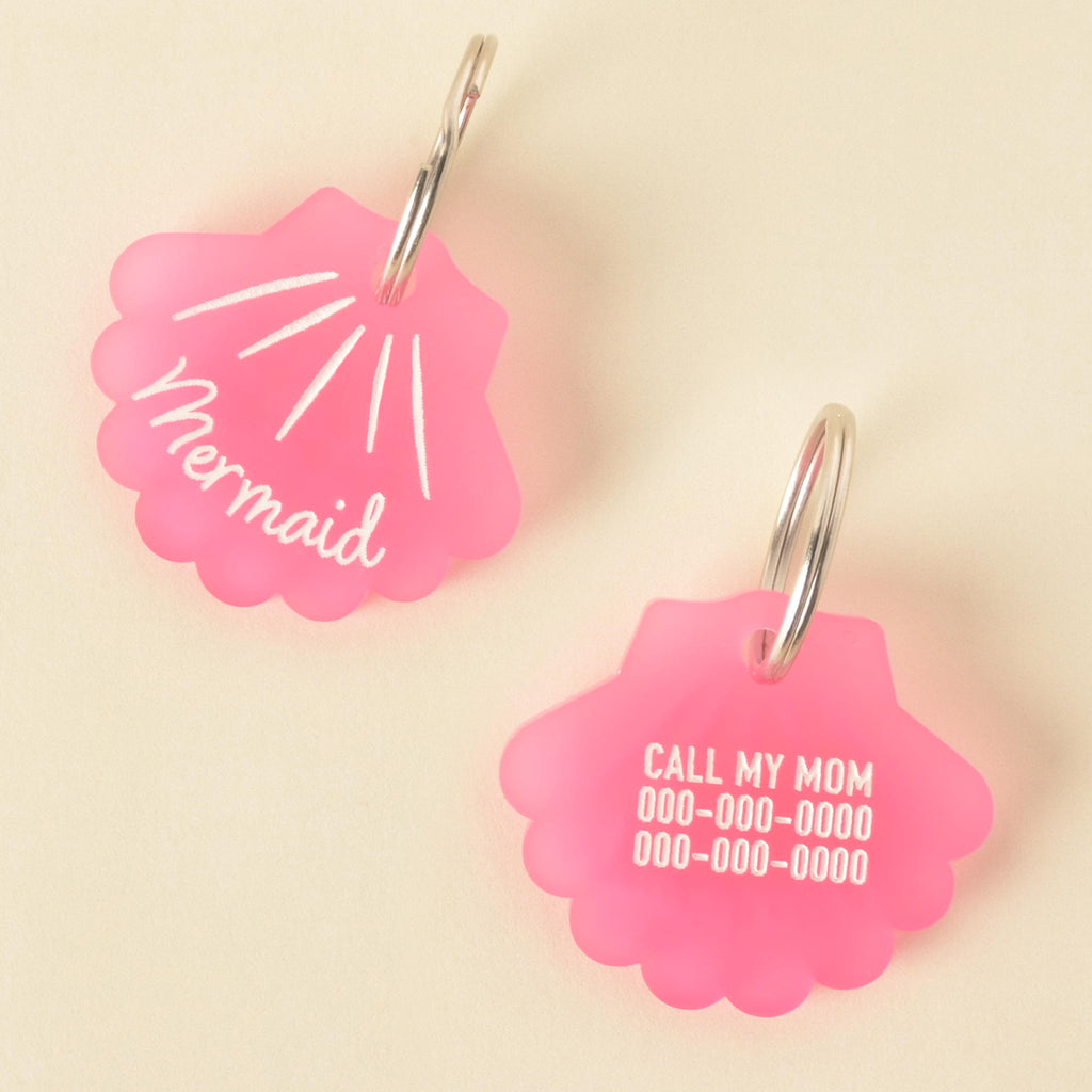pink, frosted acrylic pet ID tags shaped like a seashell. One tag is engraved with the word "Mermaid" and radiating lines, while the other is engraved with the text "CALL MY MOM" and two phone numbers. The tags are attached to silver split rings.