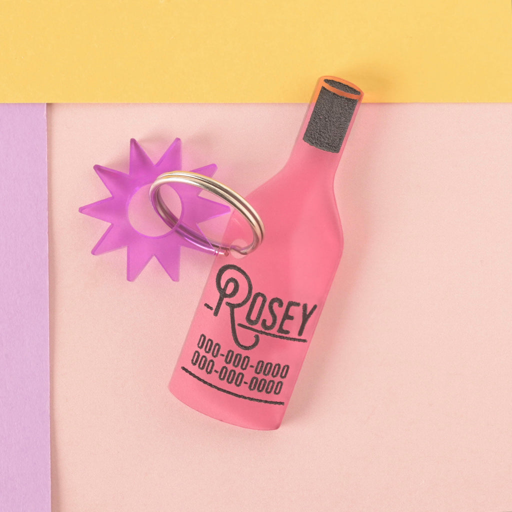 A personalized pink wine bottle-shaped pet tag with the name "ROSEY" and a placeholder phone number on it. The tag has a translucent purple starburst charm attached. The tag is connected to a silver ring for a collar and is displayed on a light pink, purple, and yellow background.
