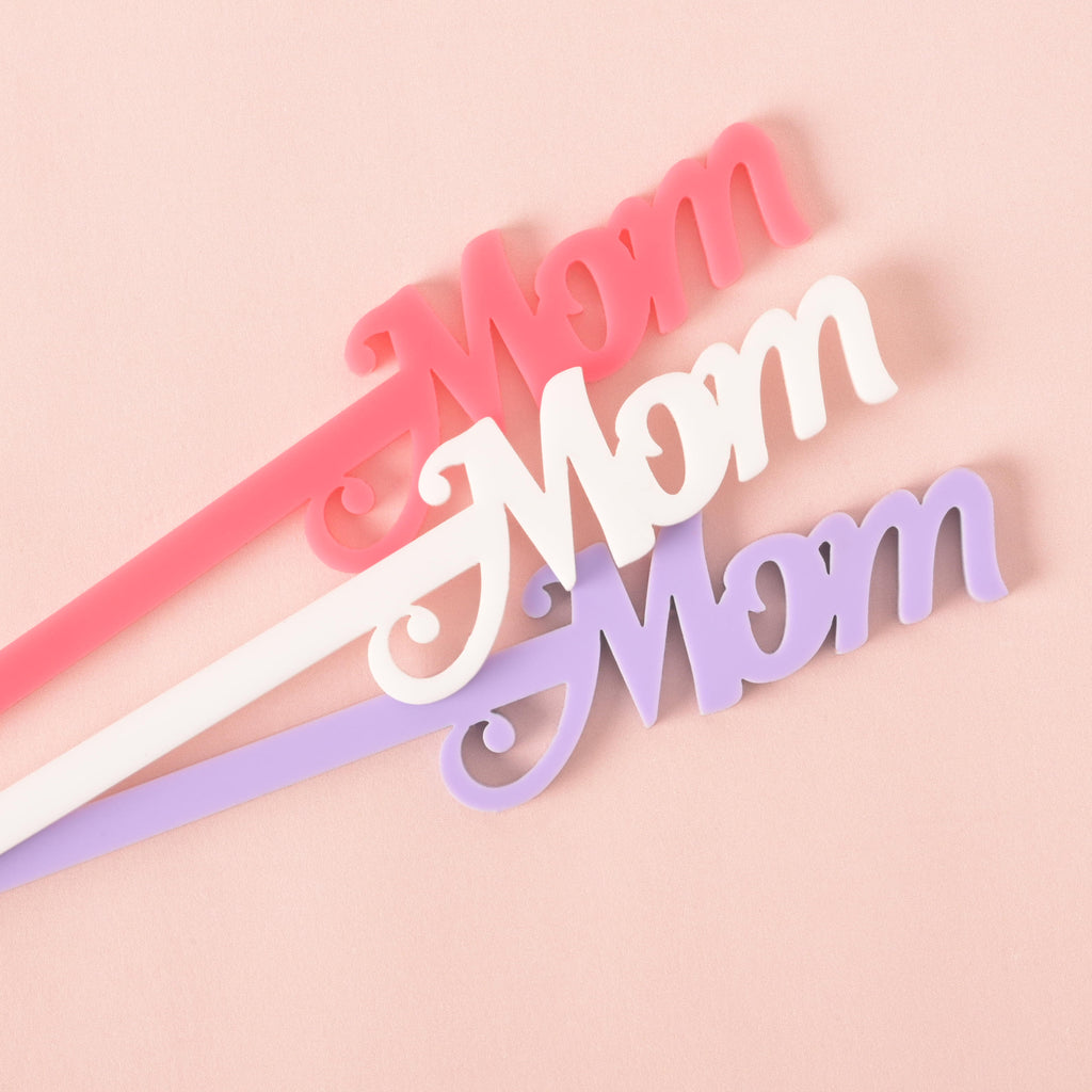 A set of three "Mom" cocktail stirrers in different colors—pink, white, and lavender. The stirrers are arranged in a fanned pattern on a solid pink background.