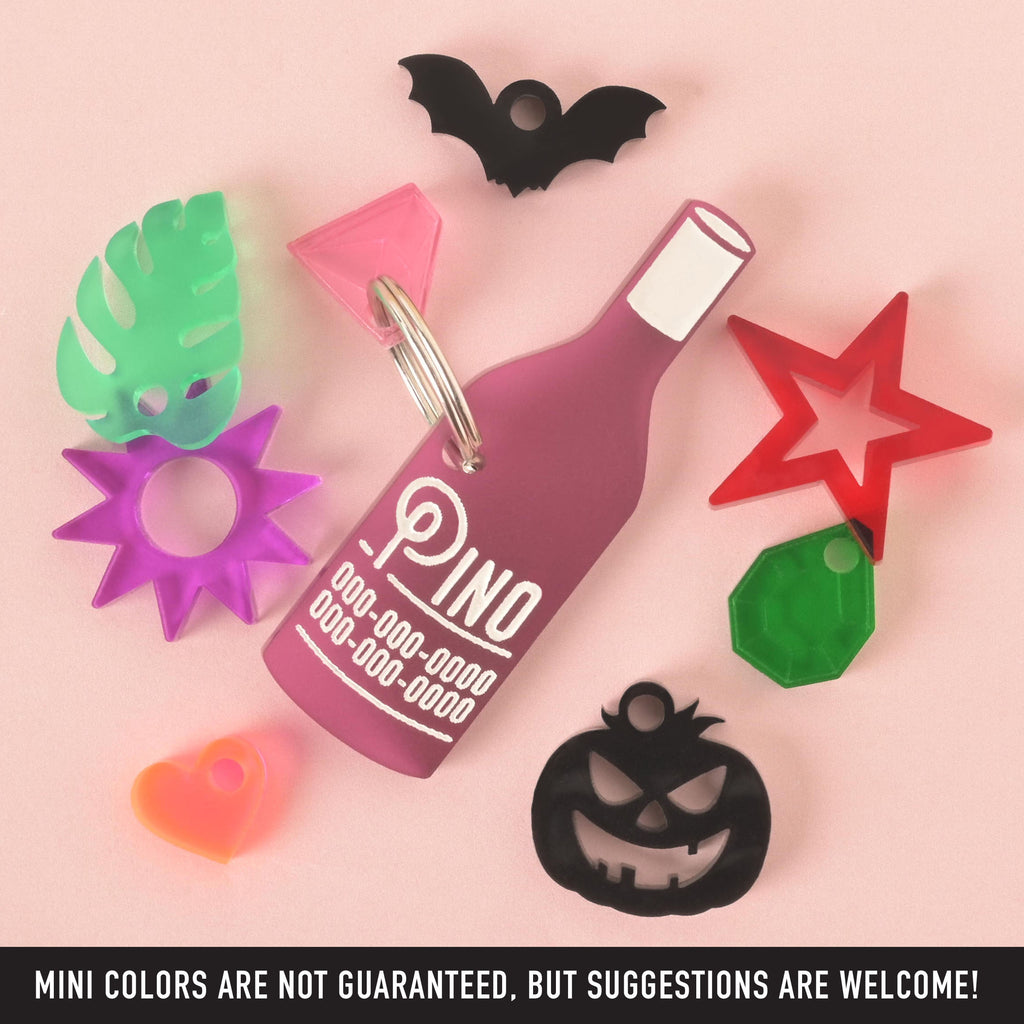 A collection of pet tags and charms scattered on a solid pink background. The central piece is a personalized pink wine bottle-shaped tag with the name "Pino" and a placeholder phone number. Surrounding it are various charms, including a green monstera leaf, a purple sunburst, an orange heart, a red star, a green gemstone, a black carved pumpkin, and a black bat. The image also contains the text "MINI COLORS ARE NOT GUARANTEED, BUT SUGGESTIONS ARE WELCOME!"