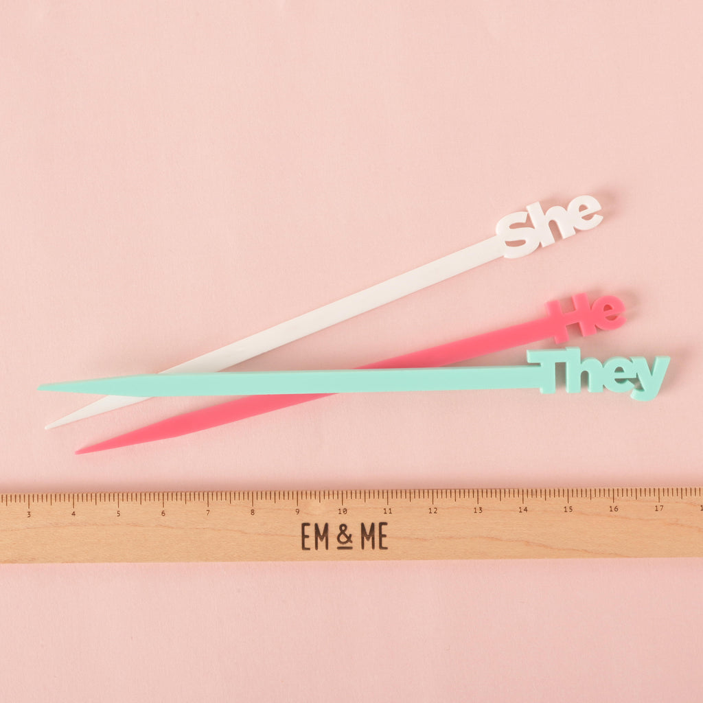 A set of three cocktail stirrers—a white "she," a pink "he," and a light blue "they"—are shown next to a ruler with the "EM & ME" logo to show their size. The background is a solid pink color.