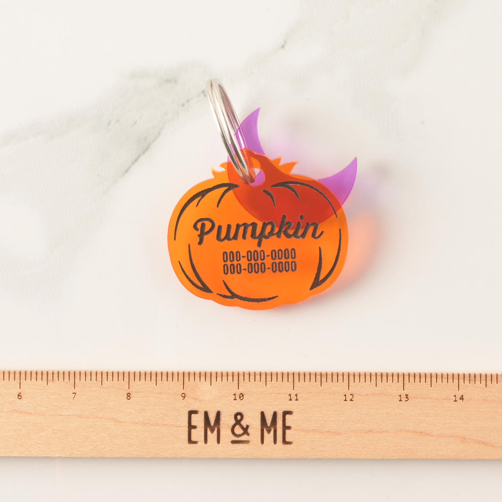 Transparent orange pumpkin-shaped pet ID tag with purple moon mini charm with a wooden ruler that reads "EM & ME" on marble background.