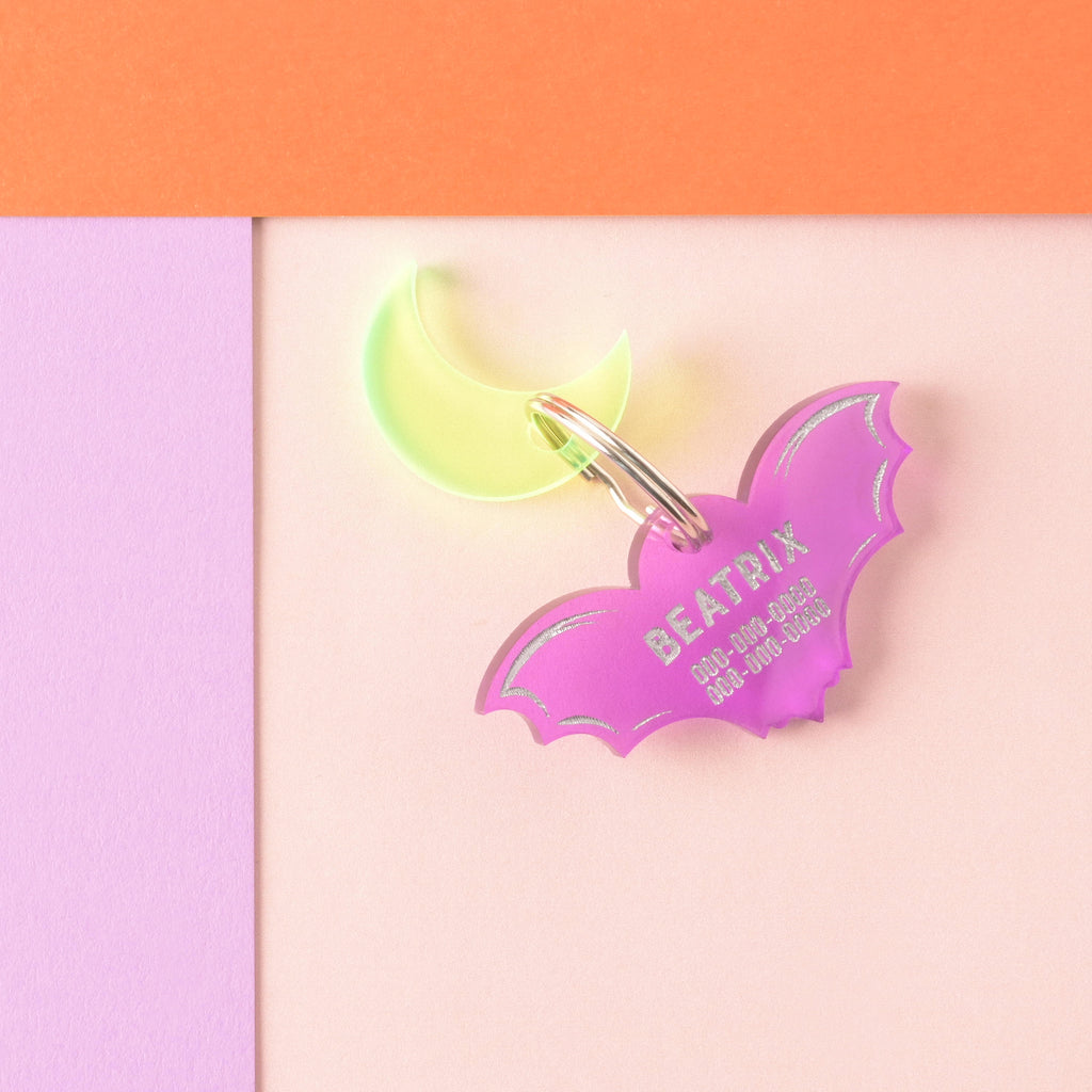 A personalized transparent purple acrylic bat-shaped pet ID tag, engraved with the name 'BEATRIX' and phone numbers in a bright silver/white. A neon yellow crescent moon charm is attached to the same ring, suggesting a celestial spooky theme.
