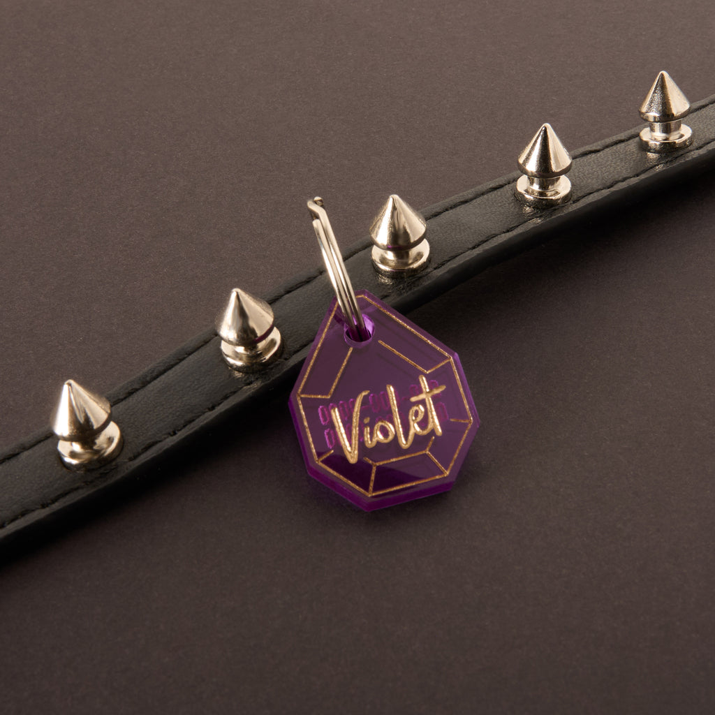 Close-up of a deep purple, faceted acrylic pet ID tag, engraved with the name 'Violet' in gold script. The tag hangs from a silver split ring against a black leather pet collar adorned with sharp silver spikes.