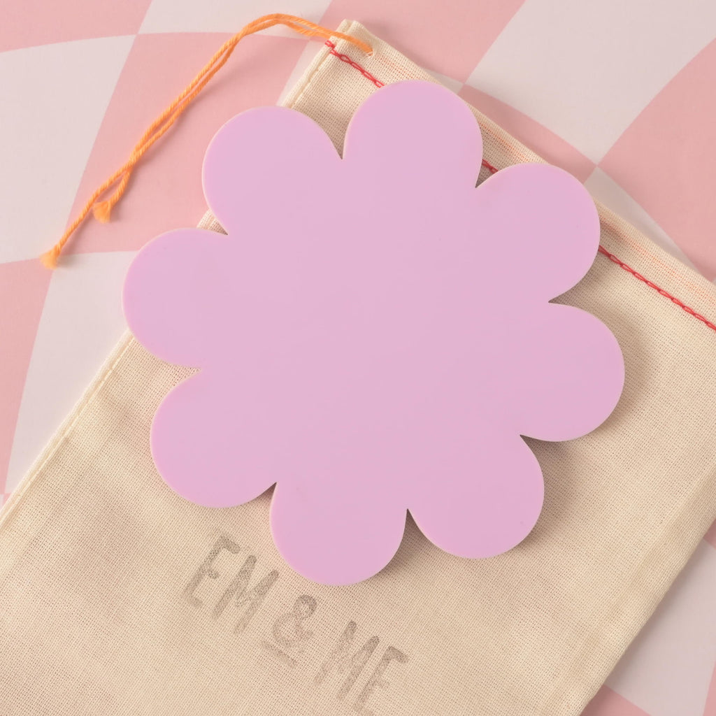 A close-up of a lilac, flower-shaped coaster , resting on a beige pouch with the faint words "EM & ME" and a pink and white checkered background.