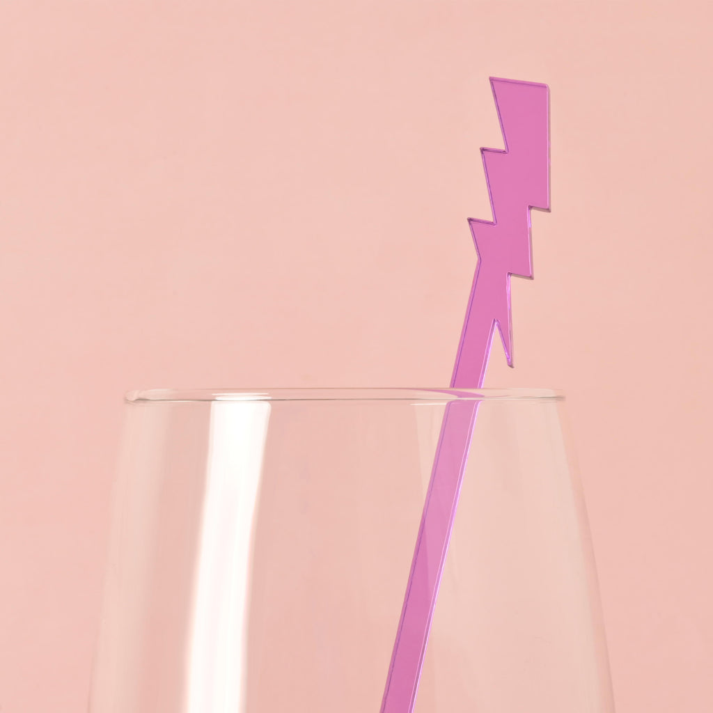 A single purple lightning bolt-shaped cocktail stirrer is placed inside a clear glass. The background is a solid pink color.