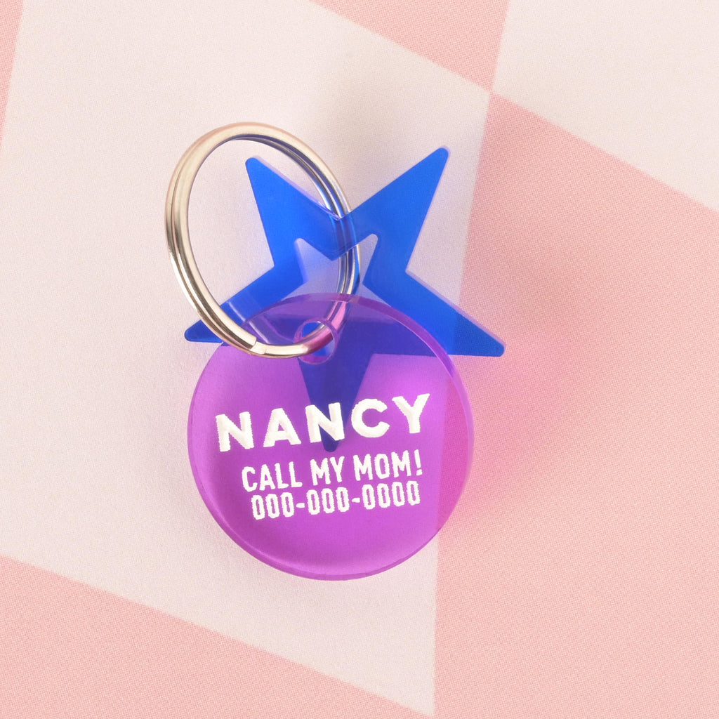 A close-up product photo of a round, purple translucent pet tag, also known as a Minimal Circle tag, with a blue star charm attached to the top. The tag is engraved with the name "NANCY" and the funny phrase "CALL MY MOM!" It is shown with a silver split key ring.
