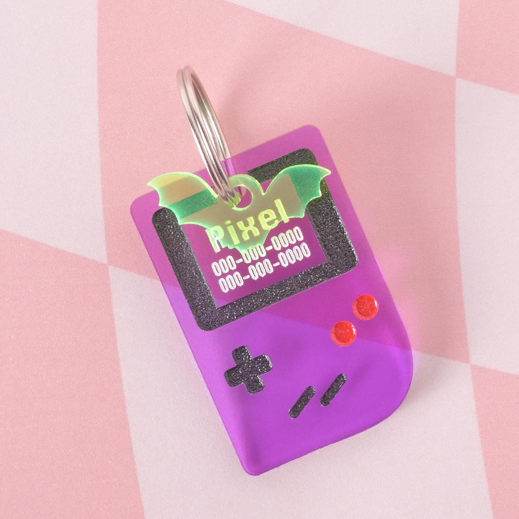 Purple retro game console pet ID tag engraved with pet name and contact number, paired with a green bat wing charm, fun and unique accessory for dogs and cats.