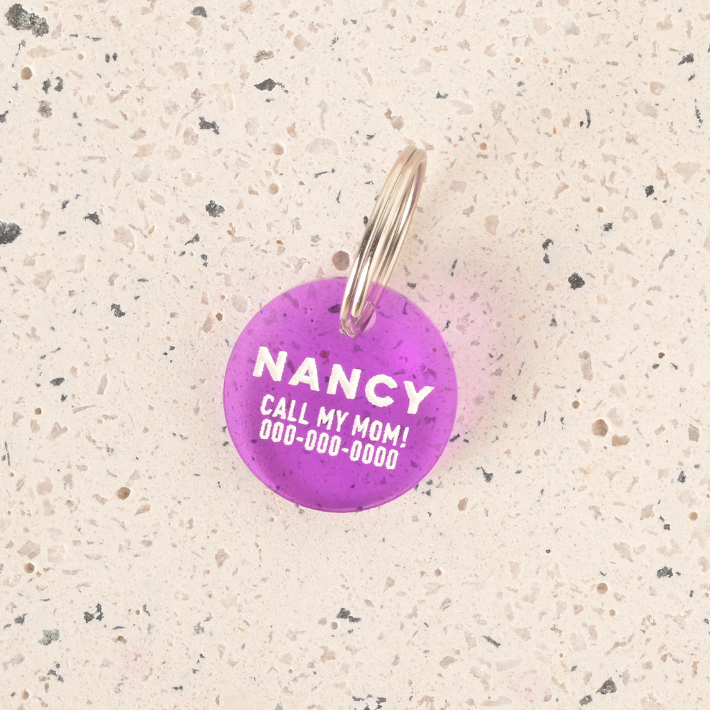 A close-up product photo of a round, translucent purple pet tag with small flecks inside. The tag is engraved with the name "NANCY" and the funny phrase "CALL MY MOM!" in white text. It is shown with a silver split key ring on a speckled beige surface.