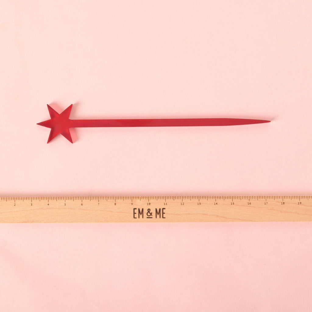 A single red cocktail stirrer topped with a six-pointed star, lying horizontally. A wooden ruler with the "EM & ME" logo is placed below the stirrer to show its length. The item is displayed on a solid pink background.