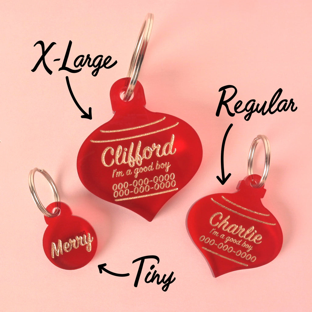 Three translucent red acrylic pet ID tags shaped like Christmas ornaments, demonstrating three sizes: 'Tiny' ('Merry'), 'Regular' ('Charlie'), and 'X-Large' ('Clifford'). All feature gold engraved holiday and personalized safety text. The tags are displayed on a light pink background.