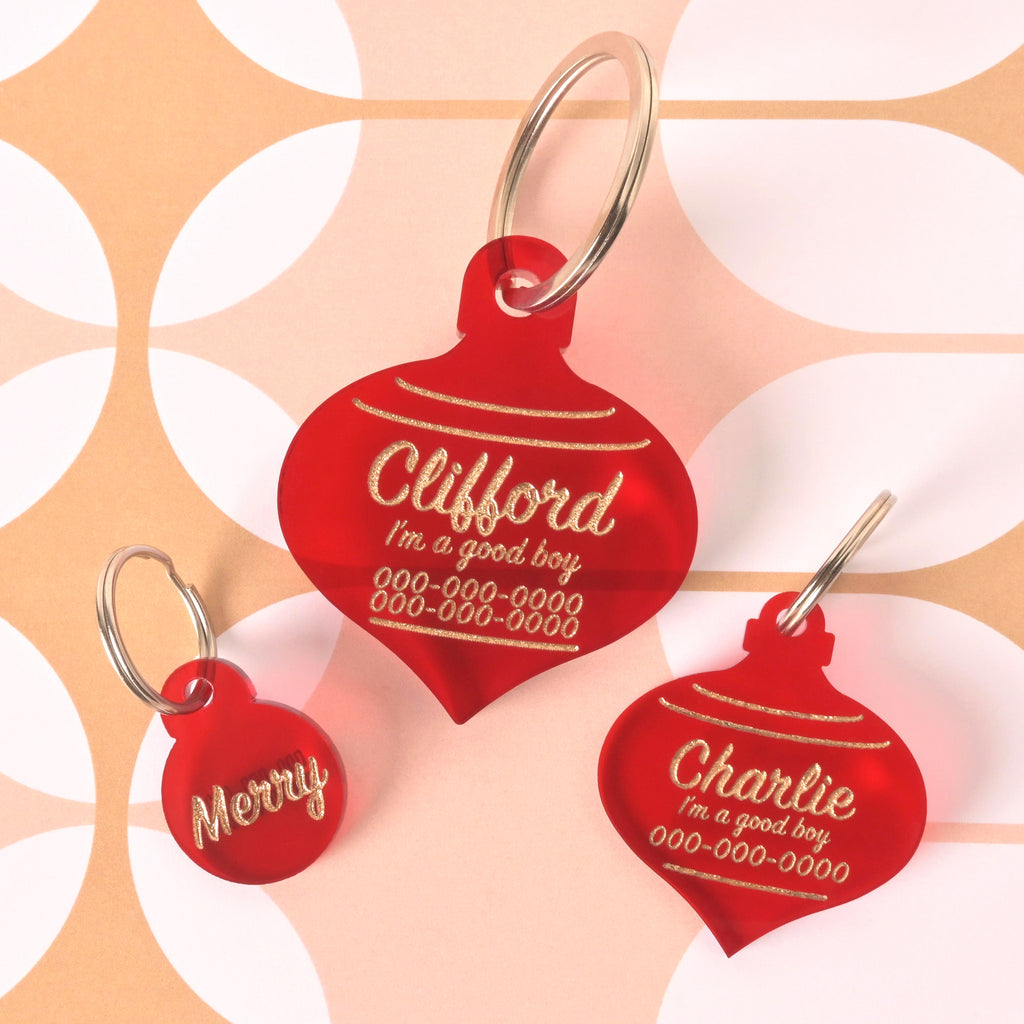 Three translucent red acrylic pet ID tags shaped like Christmas ornaments, featuring gold engraved text. The large tag is personalized for 'Clifford,' a medium tag is personalized for 'Charlie,' and a small round tag reads 'Merry.' They are displayed on a retro sunburst patterned background.