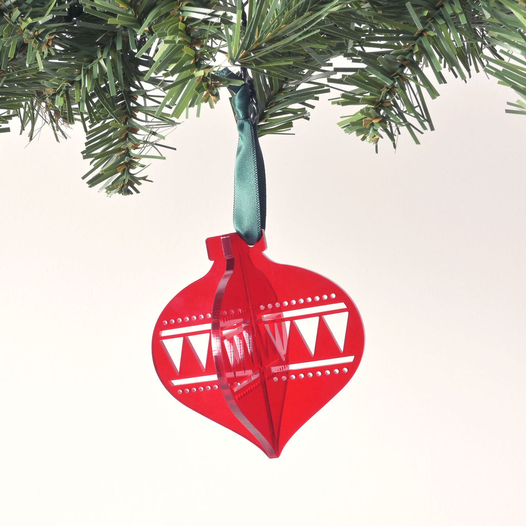 A red, flat-pack Christmas tree ornament shaped like a classic glass ornament. The assembled ornament is hanging from a pine branch with a dark black ribbon. The ornament has a zig-zag and dotted design etched on the sides. The background is a solid white color.