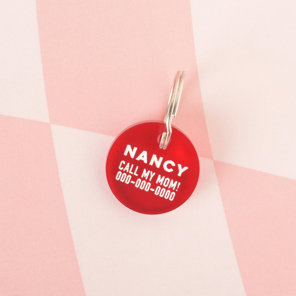 A close-up product photo of a round, red pet tag, also known as a Minimal Circle tag. The tag is engraved with the name "NANCY" and the funny phrase "CALL MY MOM!" in white text. It is shown with a silver split key ring on a checkered pink and white background.
