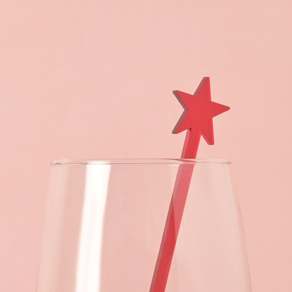 A single red star-shaped cocktail stirrer is placed inside a clear glass. The background is a solid pink color.