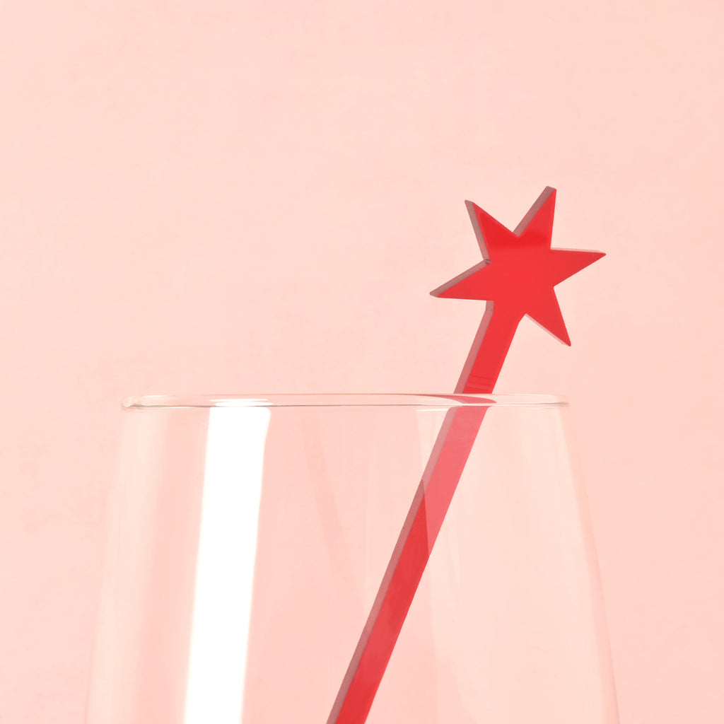 A single red Chicago star-shaped cocktail stirrer is placed inside a clear glass. The background is a solid pink color.