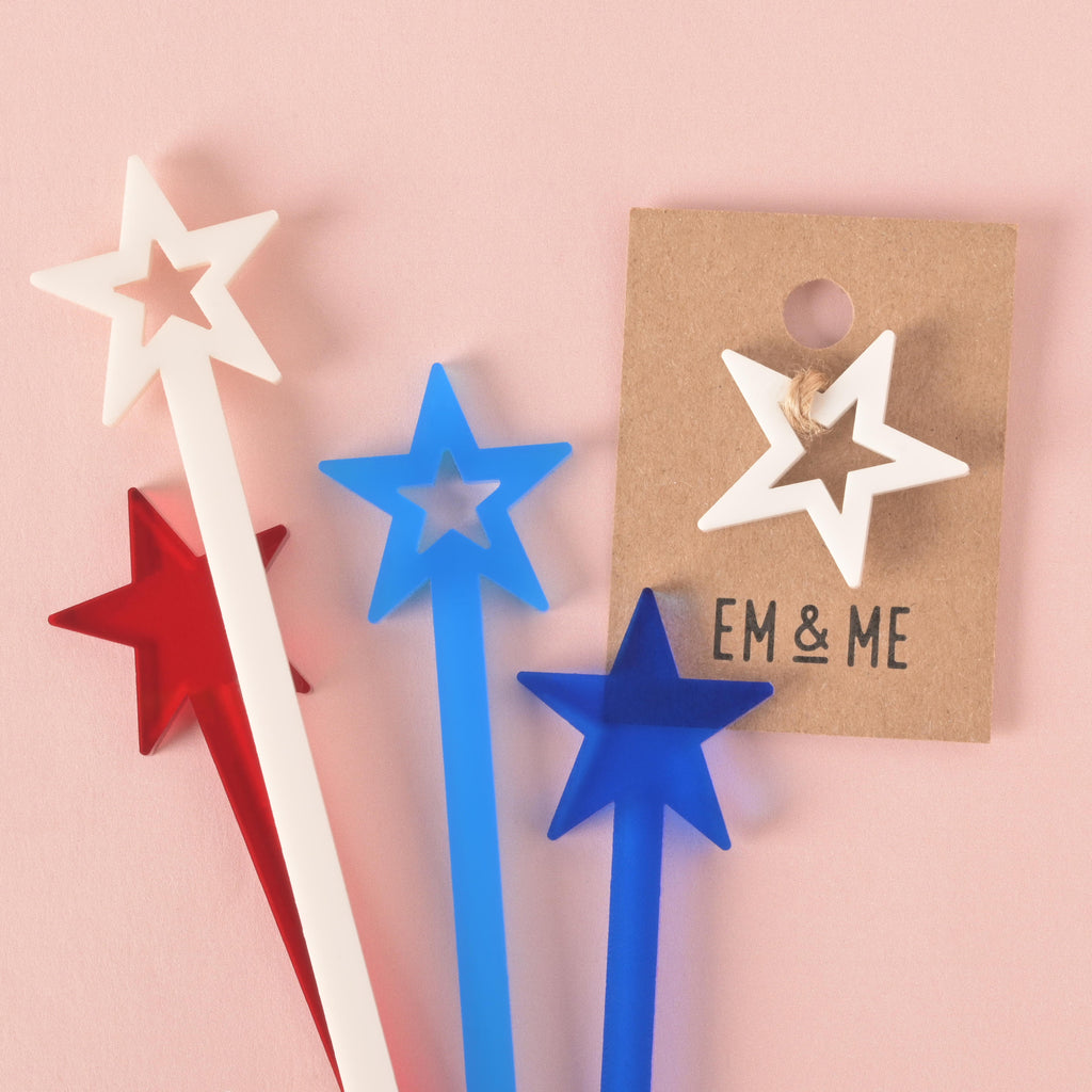 A collection of red, white, and blue star-shaped cocktail stirrers, along with a white star mini charm on a brown card. The items are displayed on a solid pink background.