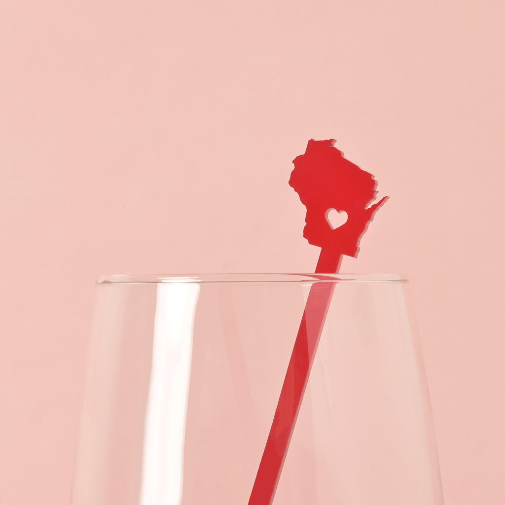A single red cocktail stirrer shaped like the state of Wisconsin with a heart cut out in the middle is placed inside a clear glass. The background is a solid pink color.